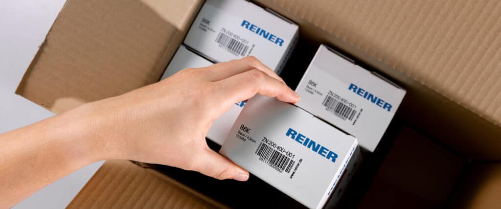 Una mano coloca un embalaje de REINER con código de barras dentro de una caja de envío.